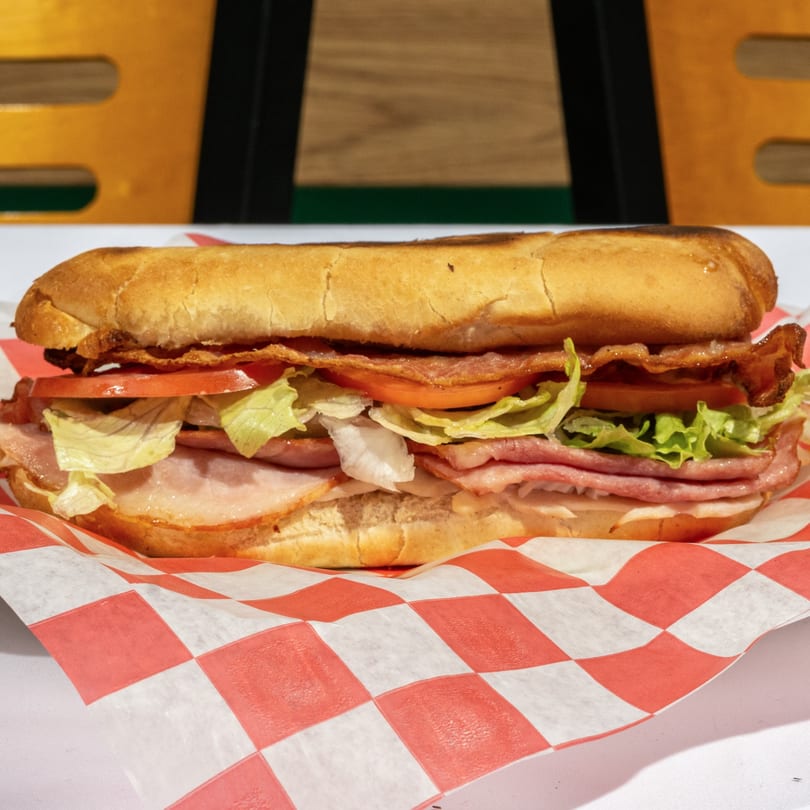 A Bear's Den Pizzeria sub sandwich with ham, bacon, lettuce, and tomato on checkered paper.